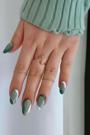 Hand with green and white nail polish wearing gold rings on a white background, Obroi