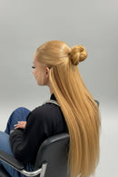 A woman shown from the side profile with long, straight blonde hair styled in a half-up bun, wearing a black salon cape against a plain light gray background, Obroi
