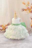 A sleeveless toddler girl's party dress displayed on a wooden mannequin. The dress features a white lace bodice, a light green satin waist sash with a bow, and a tiered ruffled skirt in white with a light green floral toile pattern, Obroi