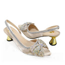 A pair of champagne satin slingback pumps with gold metallic cone heels, featuring large crystal-embellished bow details on the toes and silver rhinestone trim along the straps, shown on a white background with a 'Obroi' logo in the top-left corner, Obroi