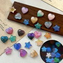 A set of small heart and star-shaped resin charms in assorted glittery and ombre colors, displayed on a wooden block and scattered on a light surface, with a circular inset showing them glowing in the dark, Obroi