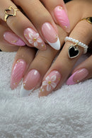 A close-up of manicured hands with long almond-shaped nails in a pink and white French tip design, decorated with 3D white daisy flowers with gold centers, small gold dots, and pearl and heart ring details, Obroi