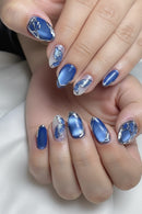 Close-up of manicured fingering with glossy blue almond-shaped nails, some featuring metallic silver glitter accents and one nail with a blue-and-white butterfly design, Obroi