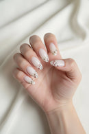 Ten false press-on nails with a glossy white base and silver French tips, decorated with small pink, gold, and silver rhinestones, arranged on a textured white fabric background, Obroi