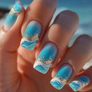 Nails with blue and white wave design and gold glitter, set against a blurred beach background, Obroi