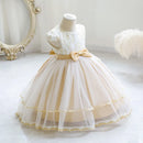 A toddler girl's formal party dress displayed on a mannequin. The dress features a cream lace bodice with a gold ribbon waistband and a layered tulle skirt in cream and gold, Obroi