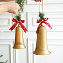 Two gold Christmas bell ornaments hanging from a white paneled wall, each decorated with a red ribbon bow, green pine sprigs, and small red berries. A hand is visible holding the left ornament's hanging string, Obroi