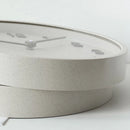 A close-up of a round wall clock with a light speckled beige face and matching base, featuring minimalist gray hands and numerals, Obroi