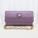 A lavender purple clutch bag covered in small rhinestones, featuring a central purple crystal brooch and a gold-tone chain strap, set against a white draped background, Obroi