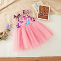 A toddler girl's sleeveless tulle dress with a pink mesh skirt and a purple floral bodice, styled on a white sheet with a beige blanket, a book, and small flowers, Obroi