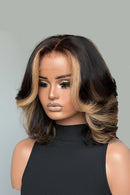 A woman wearing a short, wavy wig with dark brown roots and warm blonde highlights, styled with dramatic false eyelashes and makeup. The brand name 'Obroi' is visible in the top left corner.