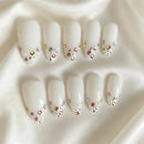 Ten white press-on nails with pointed almond tips, decorated with small silver, gold, and pink rhinestones, arranged on a draped white satin fabric, Obroi