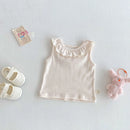 A baby girl's sleeveless top in a soft cream color with a ruffled neckline, styled in a flat lay with white baby shoes, a small pink plush bunny, and a tag on a textured light gray background, Obroi