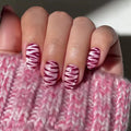 A close-up of a hand with almond-shaped nails painted in a pink and white zebra stripe pattern, resting on a chunky pink knit sweater, Obroi