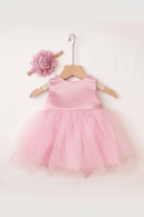 A pink sleeveless baby dress with a satin bodice and a fluffy tulle tutu skirt, displayed on a wooden hanger. The dress is styled with a matching pink rose headband and a textured pink knit blanket in the background, Obroi
