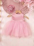A baby girl's sleeveless pink dress with a satin bodice and a fluffy tulle tutu skirt, displayed on a wooden hanger. The dress is styled with a matching pink flower headband and a pink knit hat on a soft pink background, Obroi