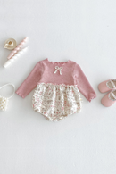 A baby girl's long-sleeve romper laid flat on a light gray background, styled with baby accessories including pink shoes, a pacifier clip, a small purse, and a teacup. The romper has a dusty pink ribbed knit top with a small bow detail, and a cream-colored ruffled underlayer with a delicate pink floral and botanical print, Obroi
