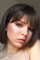 A close-up portrait of a woman with a dark brown bob haircut and straight-cut bangs, wearing a white spaghetti strap top against a neutral gray background, Obroi