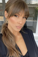 A woman with long, wavy light brown hair and straight-cut bangs, wearing a black blazer and a delicate gold necklace, photographed in a bright indoor setting, Obroi