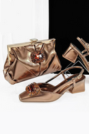 A metallic bronze evening bag and matching slingback heels displayed on a white and black studio background. The clutch bag and shoes feature a shiny satin-like finish with a jeweled rectangular embellishment on the front, Obroi