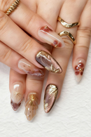 Close-up of hands with decorative nail art featuring gold and brown designs on a white background, Obroi