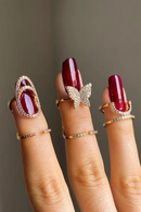 Close-up of a hand with red nail polish wearing multiple gold rings on a neutral background, Obroi