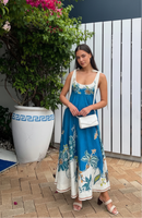 Woman in a blue floral dress standing outdoors with plants and a white wall in the background, Obroi