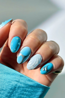 Close-up of a hand with blue and gray nail art against a blurred background, Obroi