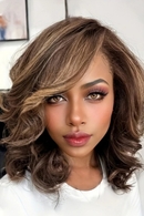 A close-up of a woman wearing a shoulder-length wavy wig with layered bangs. The wig features a dark brown base with warm caramel and light brown highlights, styled with soft curls and a side-swept fringe, Obroi