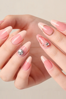 A close-up of a hand with long almond-shaped nails styled in a pink French tip manicure, featuring one silver rhinestone accent nail and a decorative pink and gold accent nail, Obroi