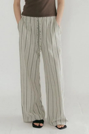 A person wearing loose, wide-leg striped pants in light beige and gray, paired with a brown top and black sandals, shown against a plain light background, Obroi