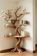 A decorative corner shelf unit shaped like a gnarled tree trunk with built-in floating shelves and faux white blossoms, styled with books, vases, and a small pitcher in a minimalist room, Obroi