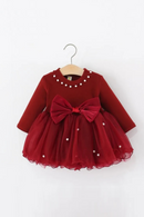 A red toddler girl's party dress with long sleeves, a large bow at the waist, and pearl embellishments, hanging on a wooden hanger against a white background, Obroi