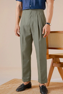 Person wearing green pleated pants and a blue shirt, standing against a beige wall with a wooden chair, Obroi