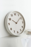 A minimalist round wall clock with a light beige face and frame, featuring dark brown Arabic numerals and matching hands, shown mounted on a white wall, Obroi